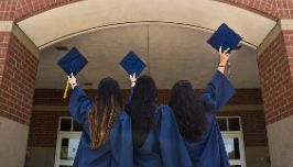  students standing in entryway wearing graduation gowns and holding their caps in their hands. 