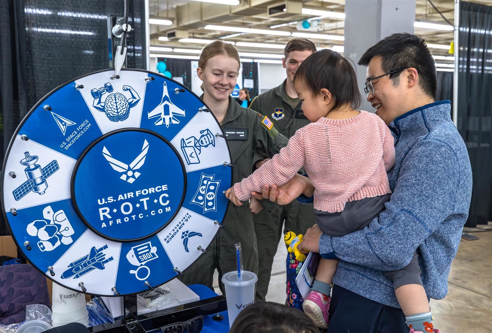 A father holds his daughter as she spins a ROTC wheel