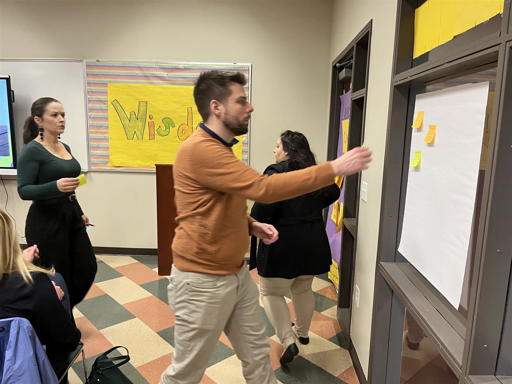 Partners walk to the front of a room to place sticky notes on paper taped to a window.